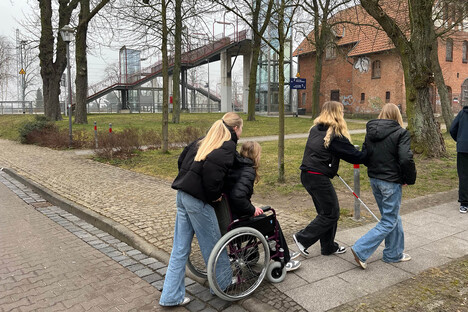 Schüler auf einem Fußweg in der Nähe einer Bahnüberführung. Eine Person sitzt im Rollstuhl eine andere hält einen Langstock.
