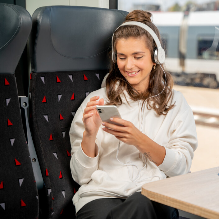 Eine Junge Frau sitzt im Zug und schaut froh auf ihr Handy.