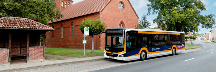 Ein Bus mit schwarzen und orangen Streifen gührt auf eine ländliche Bushaltestelle zu. Neben ihm ist ein Backsteingebäude mit einem Türmchen zu sehen
